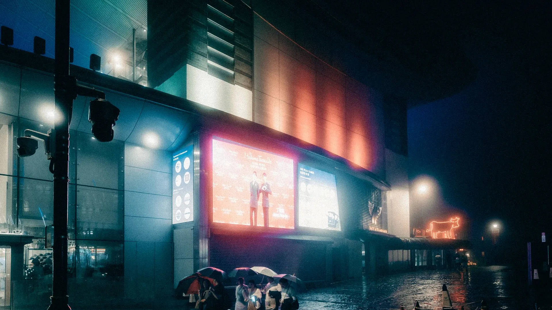 Cyan rainy street at night with people under umbrellas beneath lit billboards